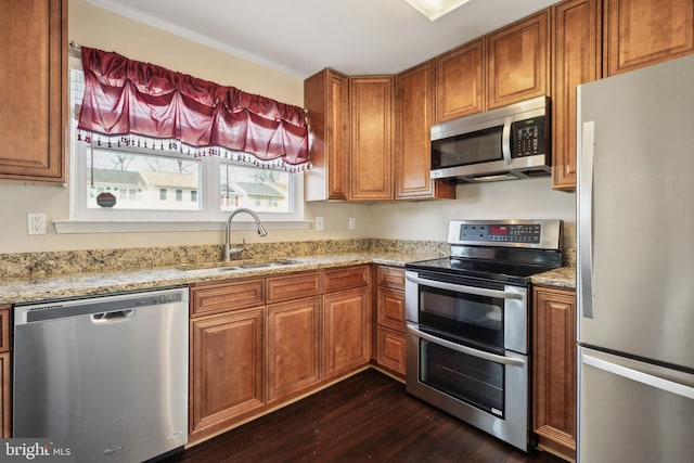 kitchen with brown cabinetry, light stone countertops, dark wood finished floors, a sink, and stainless steel appliances