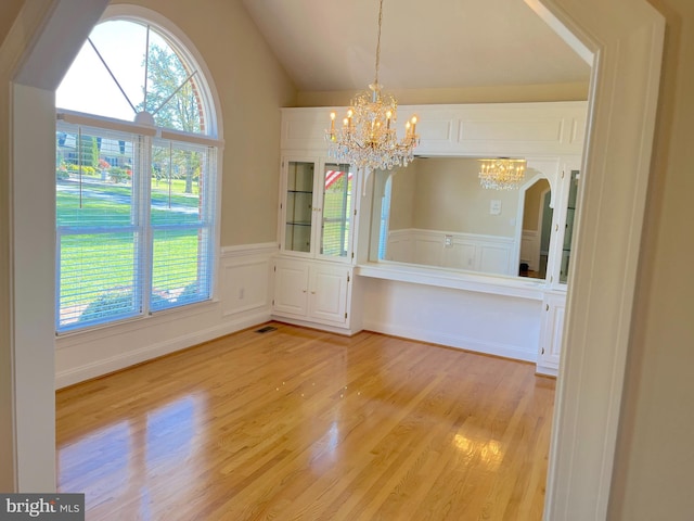 unfurnished dining area featuring light wood-style flooring, an inviting chandelier, wainscoting, a decorative wall, and vaulted ceiling