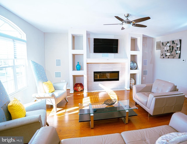 living area featuring built in features, a ceiling fan, a tiled fireplace, and wood finished floors