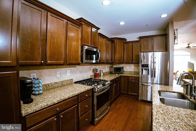 kitchen with light stone counters, stainless steel appliances, a sink, backsplash, and dark wood-style floors