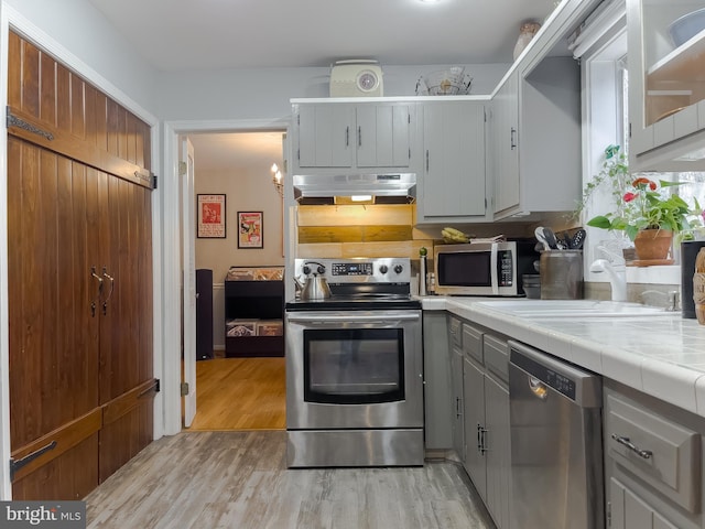 kitchen with tile countertops, light wood finished floors, a sink, stainless steel appliances, and under cabinet range hood