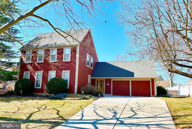 colonial-style house featuring a shingled roof, concrete driveway, an attached garage, fence, and a front lawn