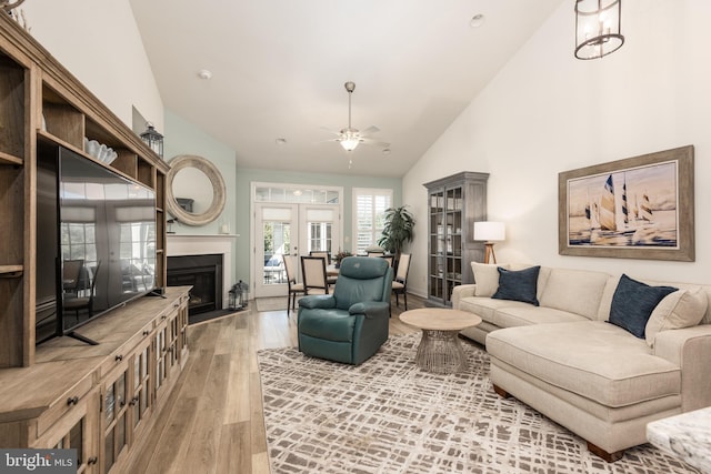 living area featuring high vaulted ceiling, light wood-style flooring, a fireplace with flush hearth, a ceiling fan, and french doors