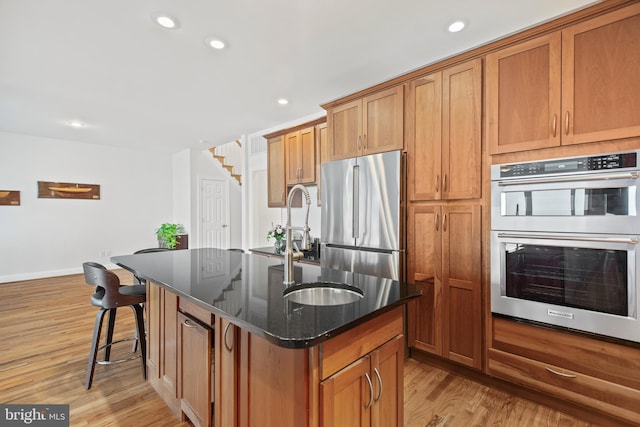 kitchen with dark stone counters, appliances with stainless steel finishes, brown cabinets, a kitchen island with sink, and light wood-style floors