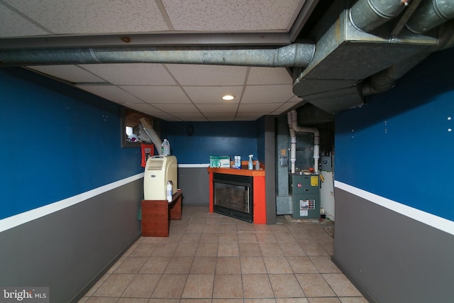 finished below grade area featuring a paneled ceiling, heating unit, tile patterned flooring, and a glass covered fireplace