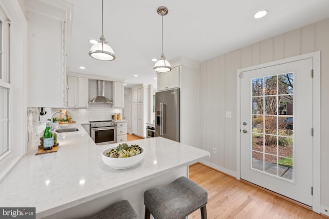 kitchen featuring light wood-style flooring, a sink, a peninsula, appliances with stainless steel finishes, and wall chimney exhaust hood