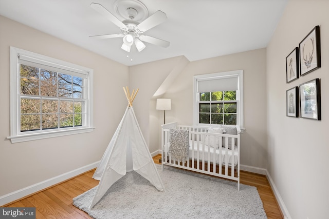 bedroom featuring ceiling fan, baseboards, a nursery area, and wood finished floors