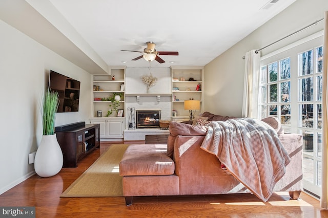 living room featuring built in shelves, wood finished floors, a ceiling fan, visible vents, and a glass covered fireplace