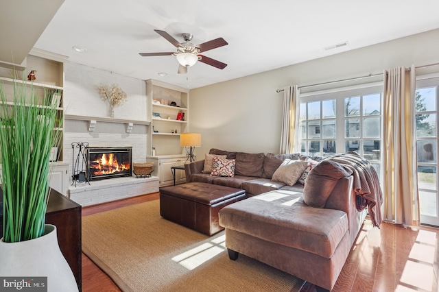 living area with ceiling fan, visible vents, a brick fireplace, and light wood-style flooring