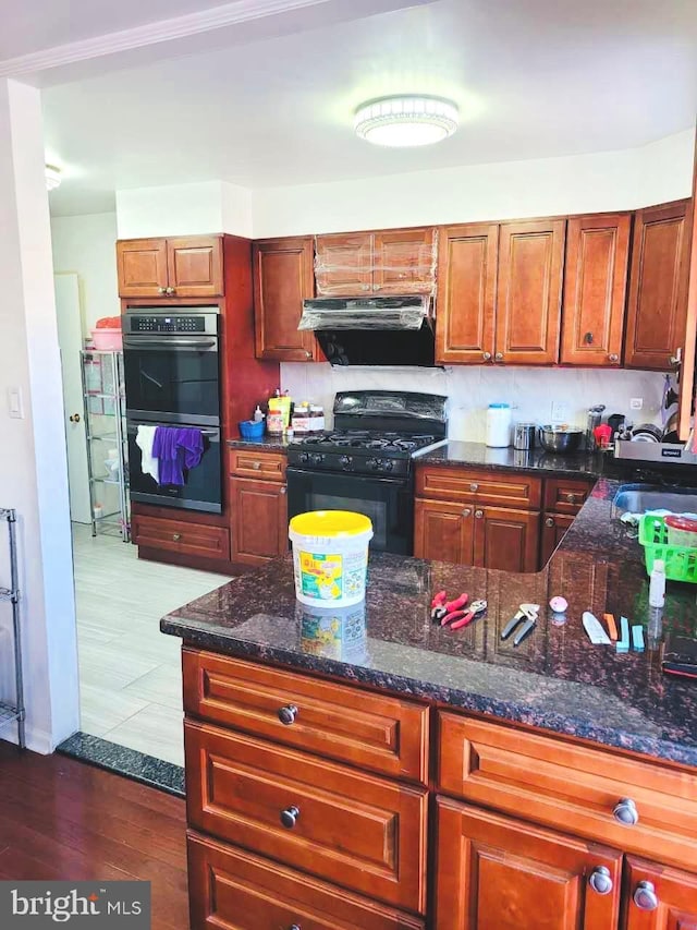 kitchen with dark stone counters, dark wood-type flooring, range hood, black gas stove, and double oven