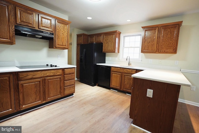 kitchen featuring light wood finished floors, a peninsula, under cabinet range hood, black appliances, and a sink