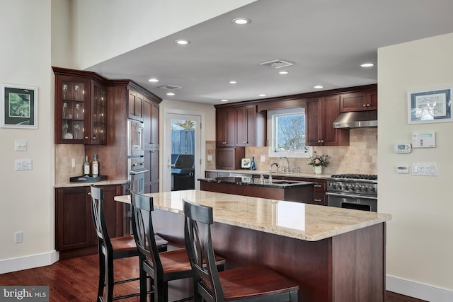 kitchen with dark wood-style floors, a center island with sink, built in microwave, stainless steel range, and under cabinet range hood