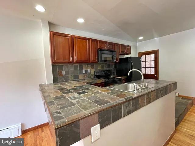 kitchen featuring light wood finished floors, tile countertops, black appliances, a baseboard heating unit, and a sink