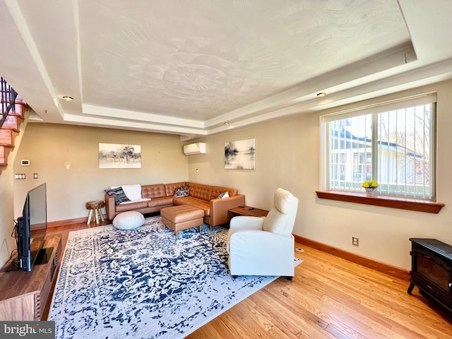 living room featuring wood finished floors, baseboards, a wall mounted AC, stairway, and a tray ceiling