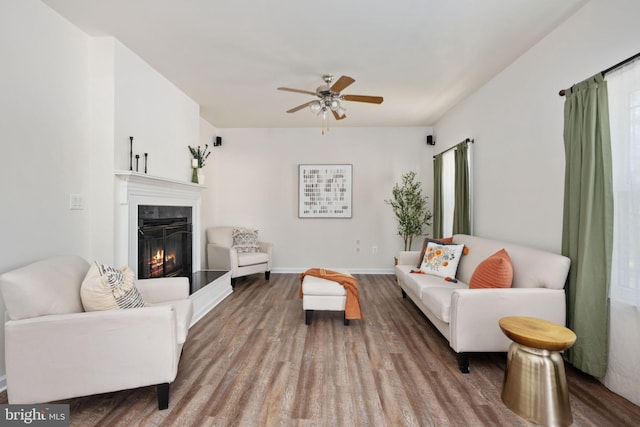 living room featuring a glass covered fireplace, ceiling fan, baseboards, and wood finished floors