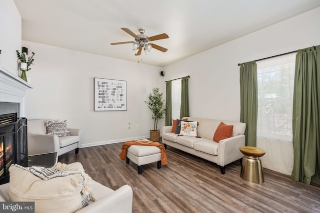 living area with baseboards, a warm lit fireplace, wood finished floors, and a ceiling fan