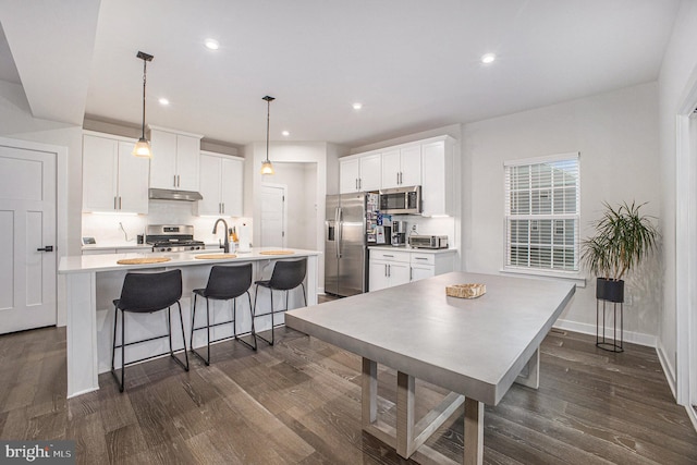 kitchen with under cabinet range hood, stainless steel appliances, backsplash, dark wood-style floors, and a center island with sink