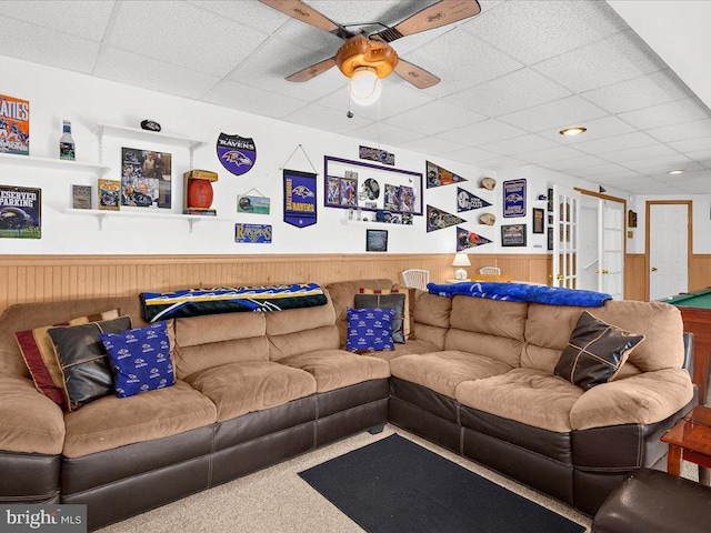 living room with ceiling fan, a drop ceiling, wood walls, and wainscoting