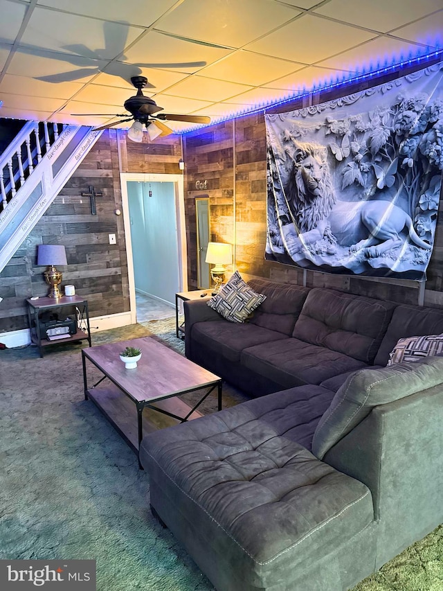 carpeted living area with wooden walls, a ceiling fan, and a drop ceiling