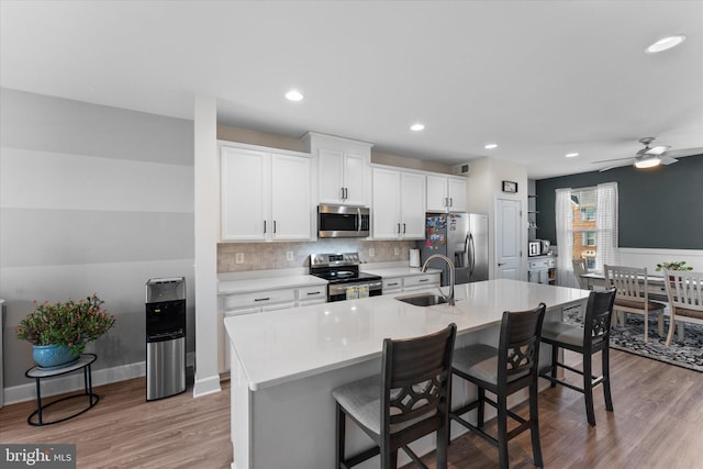 kitchen featuring light wood-style flooring, a kitchen island with sink, a sink, stainless steel appliances, and a kitchen bar