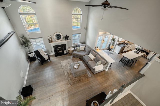 living area with hardwood / wood-style floors, a ceiling fan, baseboards, a fireplace with flush hearth, and a towering ceiling