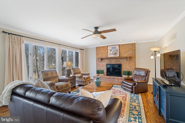 living room with visible vents, ornamental molding, wood finished floors, a brick fireplace, and ceiling fan