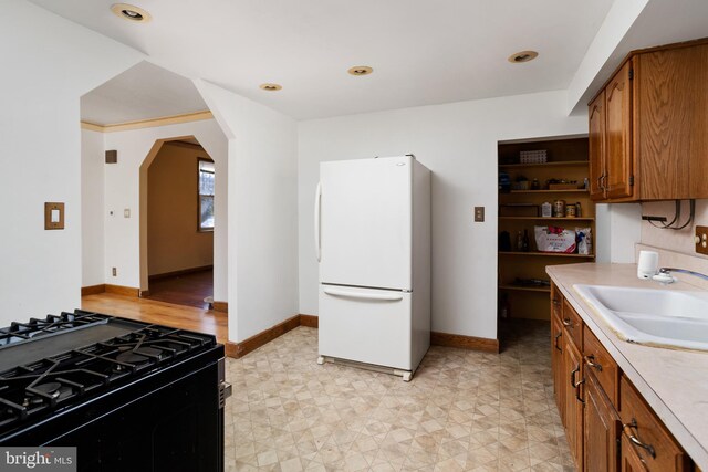 kitchen with black gas stove, brown cabinetry, freestanding refrigerator, arched walkways, and a sink