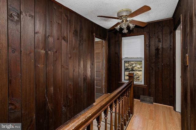 hall featuring an upstairs landing, wooden walls, light wood-style flooring, and a textured ceiling