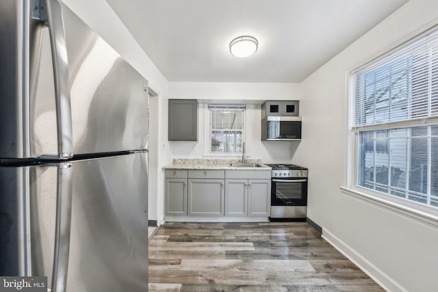 kitchen featuring wood finished floors, baseboards, a sink, gray cabinetry, and appliances with stainless steel finishes