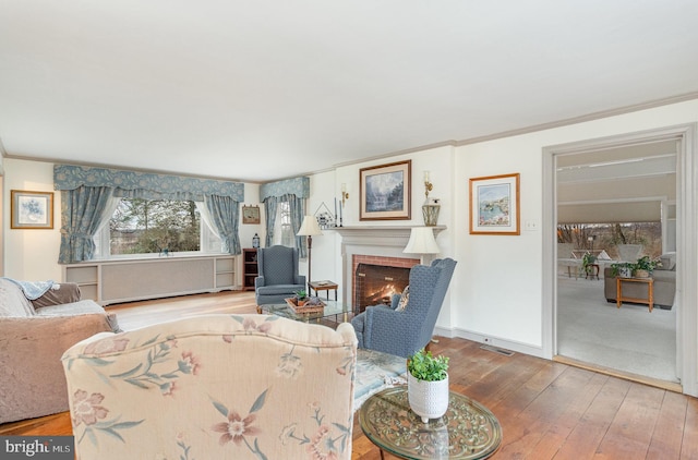 living area featuring a brick fireplace, wood-type flooring, baseboards, and ornamental molding