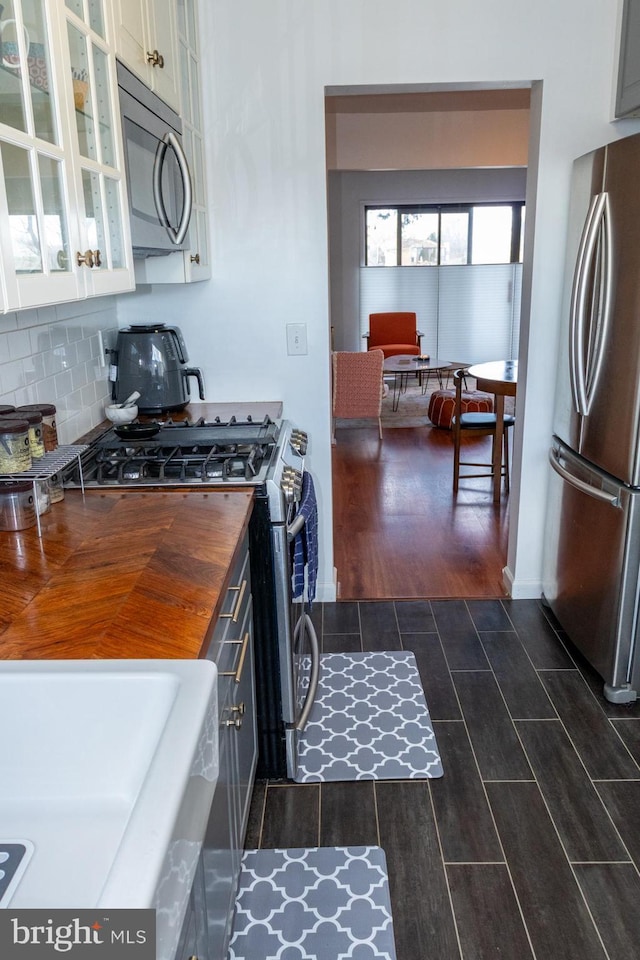 kitchen featuring stainless steel appliances, butcher block counters, backsplash, wood tiled floor, and glass insert cabinets