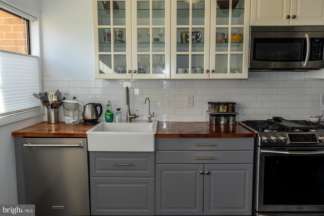 kitchen with gray cabinetry, stainless steel appliances, butcher block counters, a sink, and glass insert cabinets