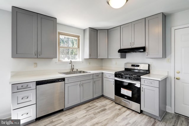 kitchen with gray cabinetry, under cabinet range hood, light wood-style floors, stainless steel appliances, and a sink