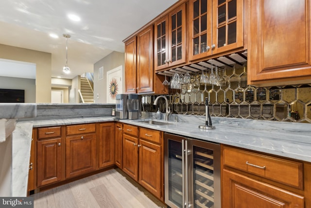 kitchen with light stone counters, brown cabinetry, a sink, wine cooler, and glass insert cabinets