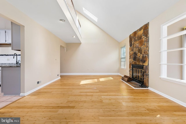 unfurnished living room with baseboards, light wood-type flooring, a stone fireplace, a skylight, and high vaulted ceiling