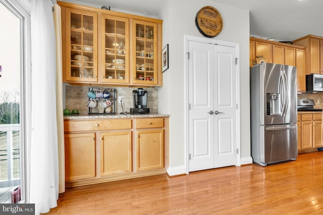 kitchen with light wood finished floors, tasteful backsplash, glass insert cabinets, light stone countertops, and appliances with stainless steel finishes