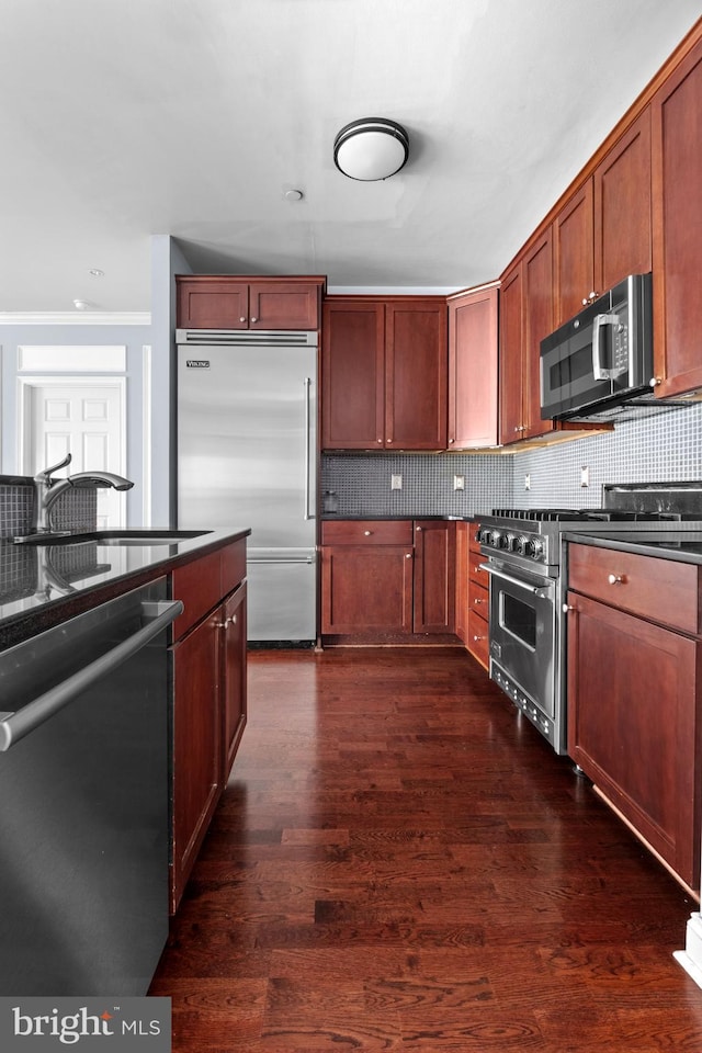 kitchen featuring dark wood-style floors, premium appliances, a sink, dark countertops, and tasteful backsplash