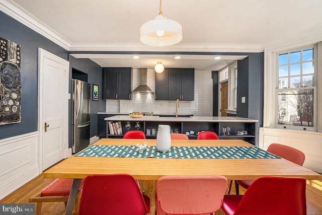 kitchen featuring a sink, dark cabinetry, freestanding refrigerator, wall chimney exhaust hood, and crown molding