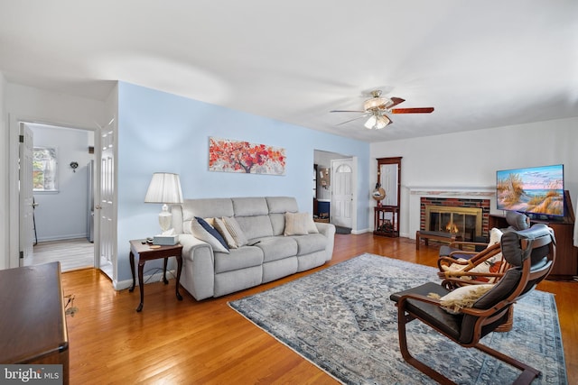 living area featuring baseboards, a brick fireplace, wood finished floors, and a ceiling fan