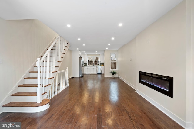 unfurnished living room featuring stairway, baseboards, dark wood finished floors, recessed lighting, and a glass covered fireplace