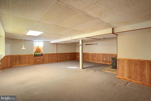 basement with a wainscoted wall, carpet, wood walls, a paneled ceiling, and a wood stove