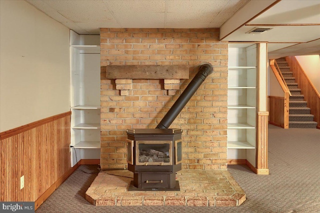 room details with wooden walls, visible vents, carpet, wainscoting, and a wood stove