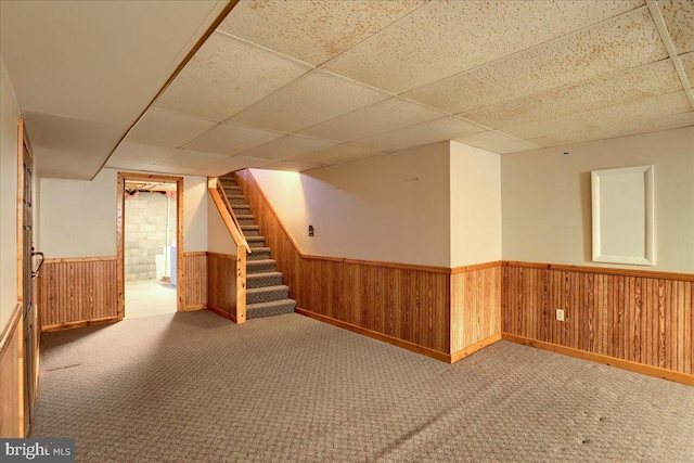 finished basement featuring stairs, wooden walls, a drop ceiling, and wainscoting