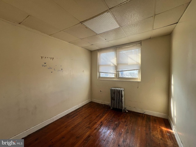 spare room featuring a drop ceiling, radiator, dark wood-type flooring, and baseboards