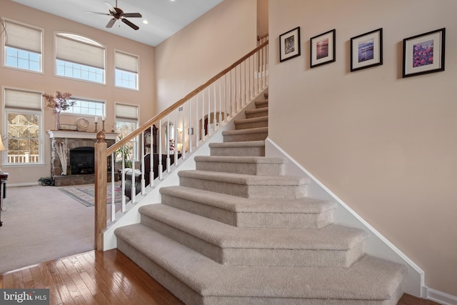 stairway featuring ceiling fan, baseboards, hardwood / wood-style floors, a fireplace, and a high ceiling