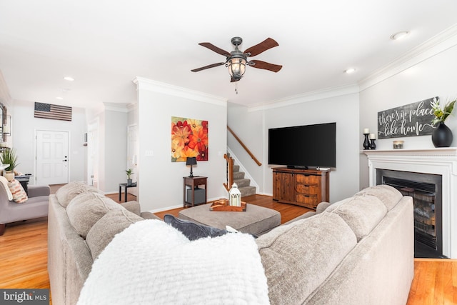 living area with crown molding, stairway, light wood-style flooring, a glass covered fireplace, and a ceiling fan