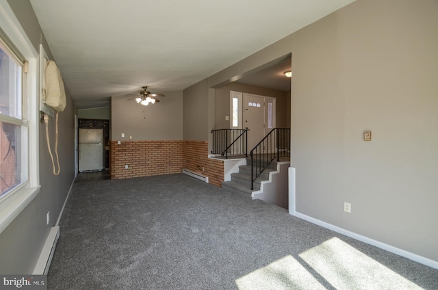 carpeted empty room with baseboards, ceiling fan, brick wall, stairs, and baseboard heating