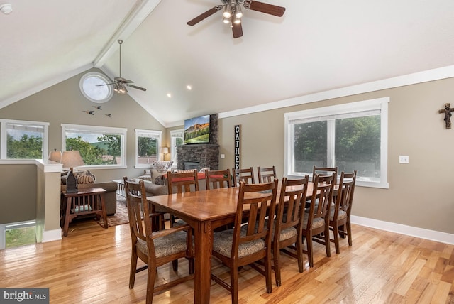 dining room with light wood-style flooring, lofted ceiling with beams, a ceiling fan, a stone fireplace, and baseboards