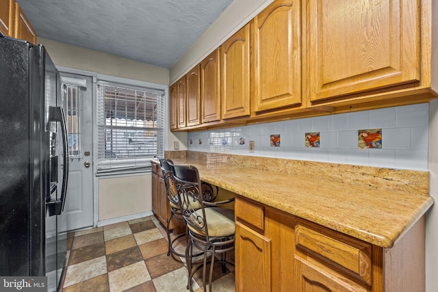 kitchen featuring brown cabinetry, built in desk, backsplash, and black refrigerator with ice dispenser