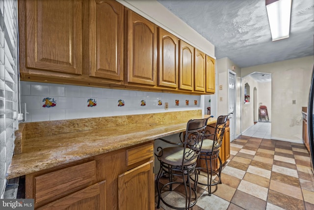 kitchen featuring decorative backsplash, arched walkways, brown cabinetry, and light stone countertops